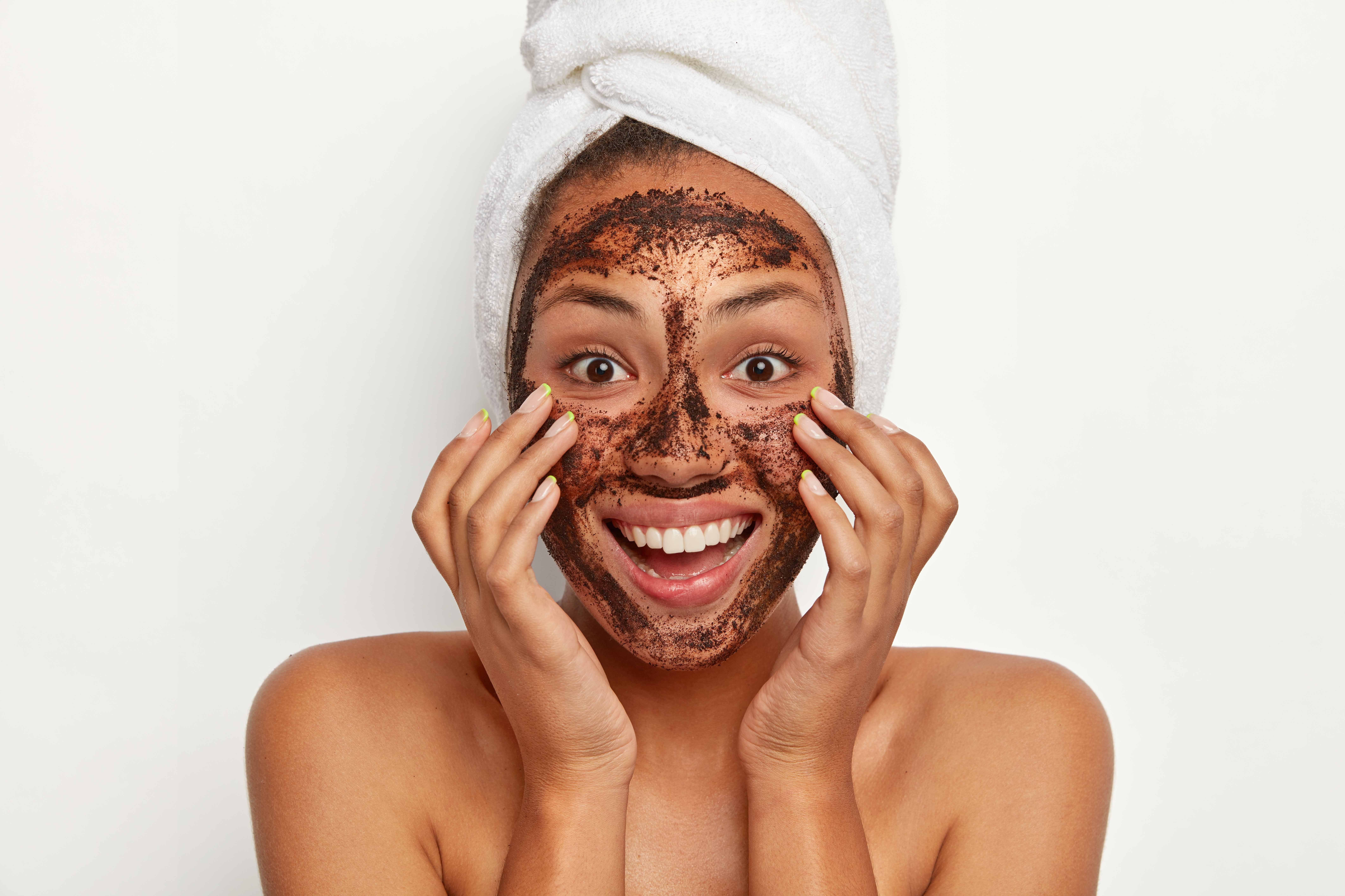 Headshot of beautiful dark skinned woman wears coffee scrub facial mask, keeps hands on cheeks, smiles broadly, wears white towel on head, models indoor against white background. Face treatment